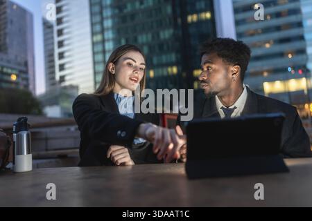 Diversi colleghi aziendali che collaborano a un progetto, analizzando i dati su un tablet digitale durante una riunione serale all'aperto in un moderno quartiere urbano Foto Stock