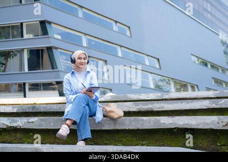 Una giovane donna asiatica che indossa cuffie e occhiali tiene un tablet mentre si trova in un parco verde vicino agli edifici del campus. Sorride con fiducia, ripreda Foto Stock