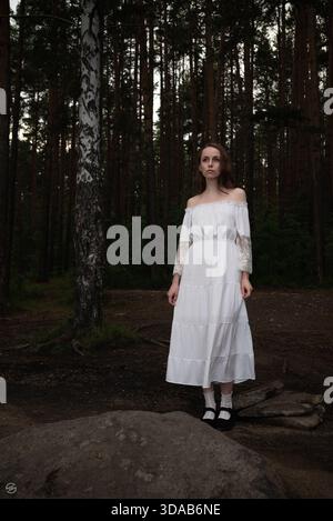 Donna in abito vintage bianco a spalla nel mezzo della foresta di pini. Foto Stock