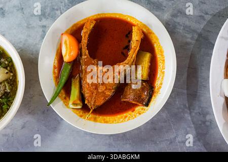 Un vivace piatto di curry di pesce con una ricca salsa rossastra, servito con pomodoro, okra, melanzane e peperoncino in una ciotola bianca. Foto Stock