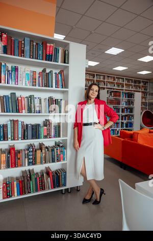 BARNAUL, RUSSIA - 7 AGOSTO. 2025: donna con capelli castani si trova in una moderna biblioteca. Indossa un vestito bianco e una giacca rossa, si appoggia su una libreria Foto Stock