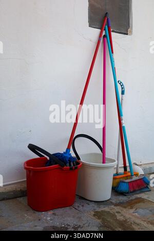 Un gruppo di utensili per la pulizia quotidiana si trova contro una parete bianca, tra cui spazzatrici, spazzoloni e secchi, ideali per lavori di sanificazione, lavori domestici e manutenzione Foto Stock