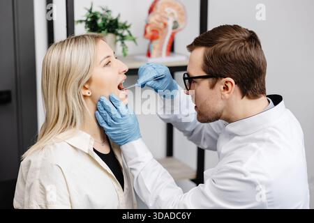 Il medico utilizza la spatola per ispezione per esaminare la gola del paziente. Medico ENT che fa esame di gola di una donna. La paziente aprì il check-up bocca-gola. Foto Stock