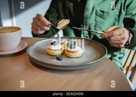 Primo piano di un uomo seduto a un tavolo che mangia Mini Pancakes con yogurt fresco, mirtilli, menta e un cappuccino Foto Stock