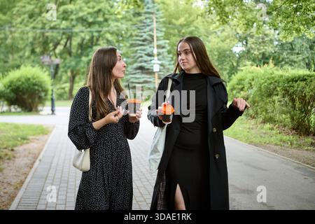 Due giovani donne con lunghi capelli castani si impegnano in vivaci pettegolezzi mentre camminano lungo il sentiero lastricato nel lussureggiante parco. La luce naturale soffusa mette in risalto il casua Foto Stock