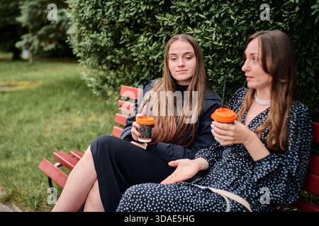 Due giovani donne sedute sulla panchina di Red Park, che tengono tazze da caffè con coperchi arancioni, si impegnano in una conversazione intima tra un lussureggiante fogliame verde, rilassandosi Foto Stock