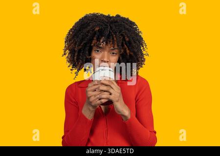 Giovane donna nera con capelli afro che tiene e beve da una tazza da viaggio su sfondo giallo Foto Stock