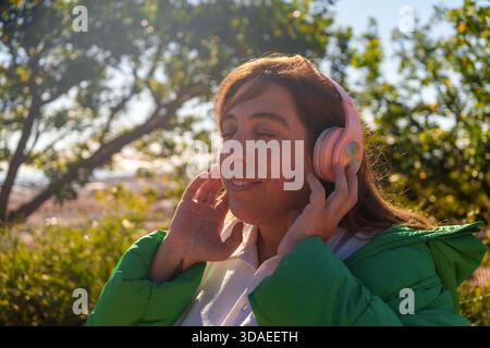 Donna che indossa cuffie rosa e giacca verde, occhi chiusi, immersa nella musica all'aperto, che sente gioia e calma sotto la luce del sole Foto Stock