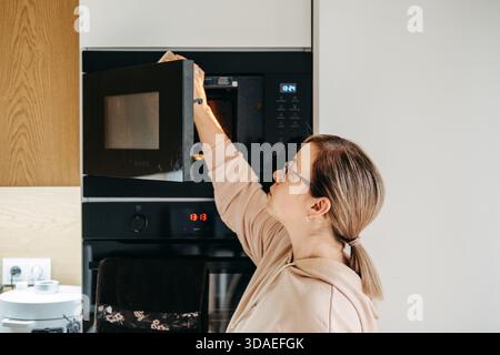 Una donna che indossa occhiali e una felpa con cappuccio entra in un forno a microonde incorporato in un moderno armadietto da cucina. Pulizia sostenibile ed ecocompatibile Foto Stock