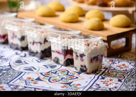 Dessert in porzioni con frutti di bosco e crema in tazze di plastica su una tovaglia a motivi a un tavolo a buffet. Primo piano di bacche e crea confezionate singolarmente Foto Stock