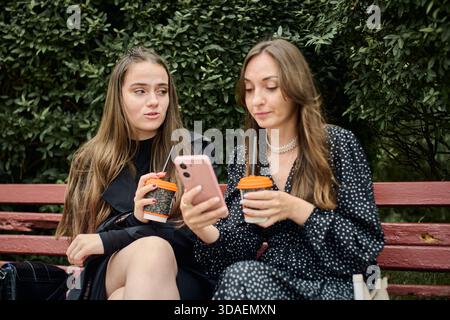 Due giovani donne con lunghi capelli marroni siedono vicino sulla panchina di Red Park, condividendo caffè e pettegolezzi guardando lo schermo del telefono, circondate dal verde folia Foto Stock