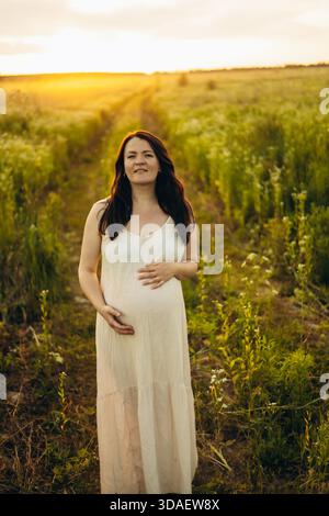 Una donna incinta cammina attraverso un campo illuminato dal sole, cullandosi la pancia e sorridendo davanti alla telecamera. Incarna serenità e anticipazione in mezzo a un verde, rura Foto Stock