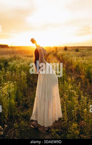 La vista laterale di una donna incinta si trova in un campo sereno, abbracciando il caldo tramonto con calma e gioia. Il suo comportamento rilassato e l'ambiente tranquillo lo trasmettono Foto Stock