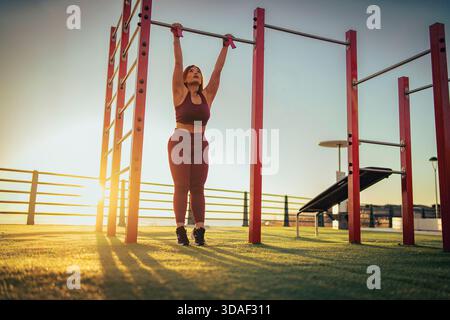 Vista posteriore di una donna sportiva appesa al bar orizzontale del parco calistenico. Silhouette di atleta che si allena all'aperto contro la luce del sole con una copia spa Foto Stock