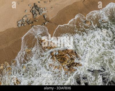 Vista aerea della costa frastagliata dove le onde schiumose si infrangono contro la spiaggia sabbiosa e gli affioramenti rocciosi, una danza di beige e bianco, Praia de Quiaios, Foto Stock