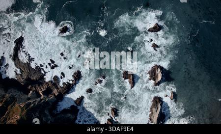 Vista aerea della costa frastagliata dove le rocce scure e frastagliate incontrano il mare turchese e le onde bianche spumeggianti si infrangono drammaticamente contro la costa, Bi Foto Stock