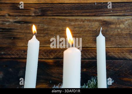 Due candele bianche bruciano con fiamme calde accanto a una terza candela spenta, tutte disposte in una corona invernale sempreverde. Foto Stock