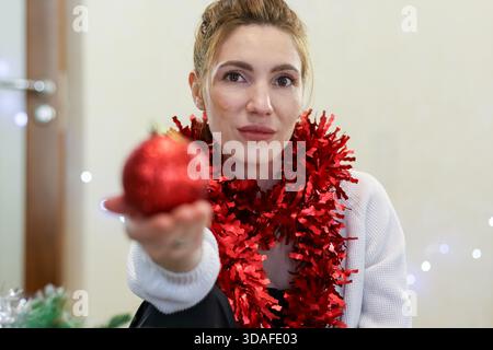 La giovane donna estende un luccicante ornamento natalizio rosso verso la fotocamera, indossando una tinsel rossa intorno al collo, circondata da alberi e luci festose. Foto Stock
