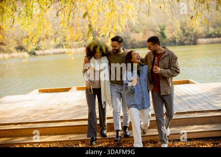 Quattro amici ridono e condividono le bevande mentre camminano lungo un sentiero di legno vicino a un tranquillo lago circondato da alberi autunnali. Foto Stock