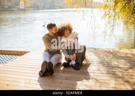 Due persone siedono a stretto contatto su un molo di legno, sorridendo e condividendo un momento tenero sotto la calda luce del sole sul lago. Foto Stock