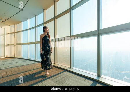 Bella donna asiatica elegante in un elegante abito in piedi davanti a una finestra di vetro all'interno di un moderno edificio alto con lo skyline della città di Bangkok Foto Stock