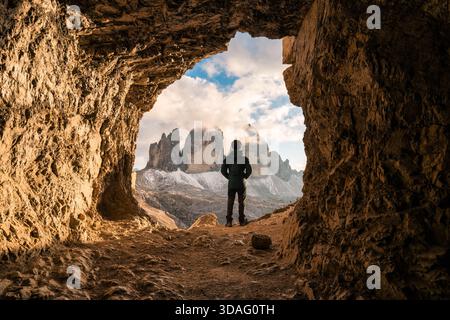Vista posteriore del viaggiatore maschile in piedi di fronte alla grotta rocciosa Grotta delle tre Cime con picco di montagna durante la serata nelle Dolomiti, Italia Foto Stock