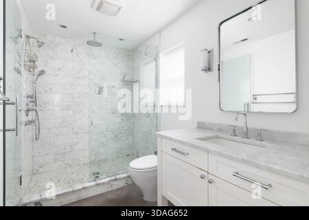 Un bagno bianco luminoso e moderno caratterizzato da una spaziosa cabina doccia in vetro con piastrelle in marmo e un piano lavabo bianco con ripiano in marmo. Foto Stock