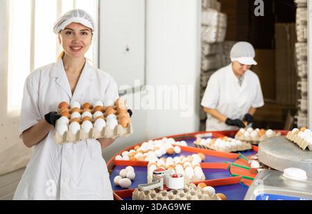 Felice giovane agricoltore europeo in uniforme che detiene una scatola di cartone piena di uova di pollo fresche dopo la cernita e l'etichettatura nell'azienda avicola Foto Stock