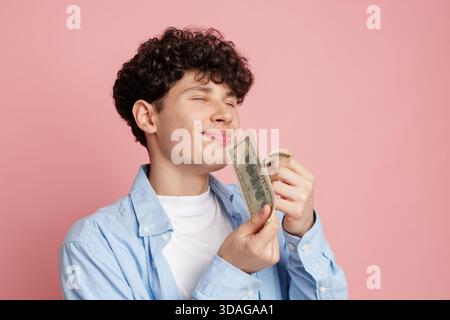 Il giovane tiene le banconote vicino alla faccia con un'espressione soddisfatta che mostra il desiderio di denaro l'avidità la motivazione finanziaria e lo stile di vita ricco in studio Foto Stock