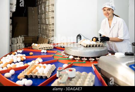 Giovane agricoltrice positiva con cappotto bianco che ordina in base alla taglia e segna le uova di pollo fresche sul nastro trasportatore dalla penna con i polli Foto Stock