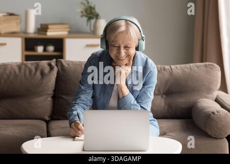 Donna anziana con auricolari che studia utilizzando un notebook e prende appunti di carta Foto Stock