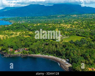 Vista aerea in droni della costa di Tulamben con spiaggia di sabbia nera, barche e montagne Foto Stock