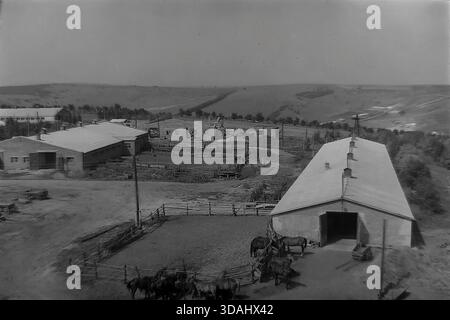 Bohorodychne, regione di Donetsk, RSS Ucraina, anni 1970 Una vista panoramica archivistica di un grande complesso agrituristico situato in una valle vicino alle colline. Lunghe stalle in mattoni e stalle con tetti in ardesia sono disposte intorno a cortili di sterrato. In primo piano, i cavalli sono visti in coralli di legno, mentre i materiali da costruzione suggeriscono lo sviluppo continuo delle infrastrutture rurali. Questo paesaggio cattura le dimensioni dell'allevamento e l'atmosfera tranquilla della campagna sovietica nel Donbas. Foto Stock