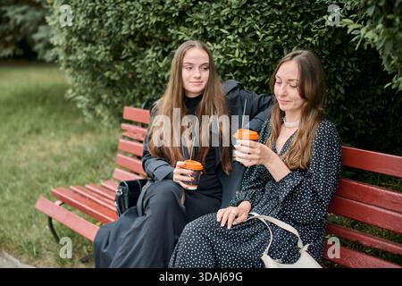 Due giovani donne con lunghi capelli marroni siedono sulla panchina di legno rossa all'esterno, circondate da verde vegetale, bevono caffè e condividono momenti di relax insieme Foto Stock