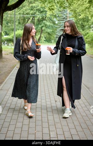 Due giovani donne con lunghi capelli scuri si impegnano in una vivace conversazione tenendo in mano tazze da caffè, camminando sul sentiero lastricato nel lussureggiante parco verde durante il giorno Foto Stock