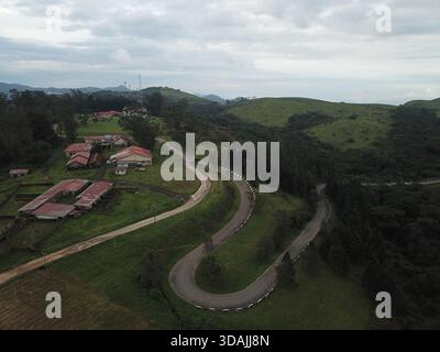 Vista aerea di una strada tortuosa che serpeggia attraverso lussureggianti colline e valli verdi, in contrasto con gli edifici dal tetto rosso annidati nel verde, Obudu Foto Stock