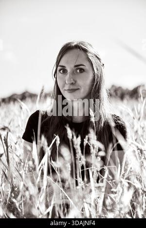 Ritratto di una giovane donna con capelli lunghi in piedi tra alti gambi di segale in piena luce del giorno. Lo sfondo morbido e l'ambiente naturale sottolineano la calma e la serenità Foto Stock