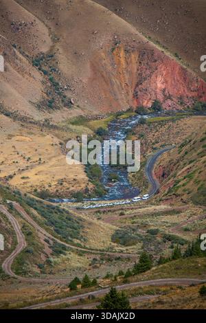 Vista ad alto angolo di una linea di veicoli fuoristrada che viaggiano lungo una tortuosa strada sterrata accanto a un fiume in un aspro canyon di montagna. Foto Stock