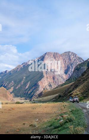 Regione di Naryn, Kirghizistan - 13 maggio 2024: Un convoglio di veicoli da spedizione attraversa una valle verso massicce vette rocciose rosse Foto Stock