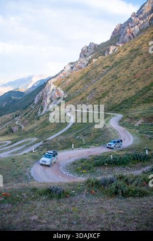 Regione di Naryn, Kirghizistan - 13 maggio 2024: Auto che guidano su una strada di montagna ghiaiosa con molte curve che si snodano attraverso ripidi pendii erbosi Foto Stock