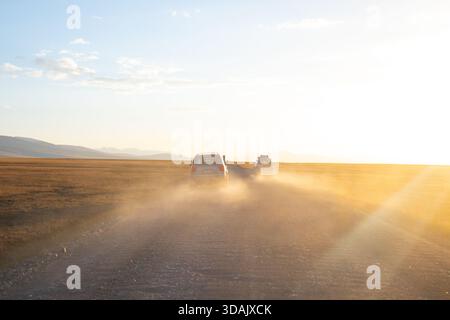 Regione di Naryn, Kirghizistan - 13 maggio 2024: I veicoli sollevano polvere pesante mentre guidano velocemente su una strada sterrata verso il sole che tramonta Foto Stock