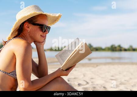 Donna spensierata che indossa un cappello da sole che si rilassa sulla spiaggia leggendo il libro. Foto Stock