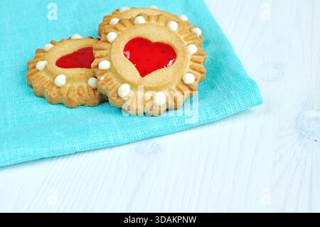 Concetto di pasto romantico - tre biscotti di pasta frolla al burro con gelatina a forma di cuore rosso sulla tovaglia di lino turchese sopra il tavolo di legno bianco. Fr Foto Stock