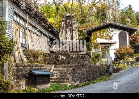 Una pietra commemorativa recante un'iscrizione giapponese su una scala di pietra alla periferia di Tsumago-juku, con tradizionali edifici in legno e cemento e fogliame autunnale sullo sfondo, Nagiso, prefettura di Nagano, Giappone Foto Stock