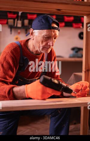 Professione, arte e concetto di hobby. Carpentiere uomo anziano che indossa tute blu per assemblare scaffali in legno utilizzando trapani elettrici che lavorano su legno in garage. Foto Stock