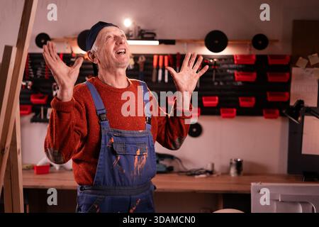 Arrabbiato aggressivo falegname maturo urla con le mani alzate a causa di problemi aziendali in officina. Fatti a mano, piccole imprese, fai da te e frustrazione Foto Stock