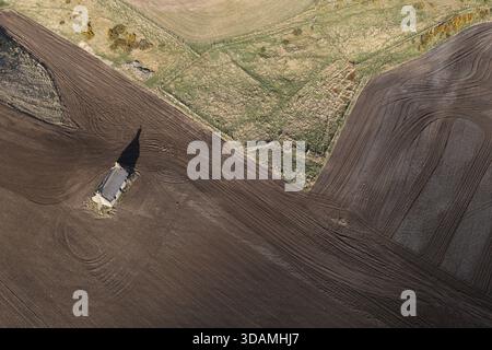 Veduta aerea di un edificio solitario che getta una lunga ombra attraverso la terra scura appena lavorata e incontra i campi verdeggianti in un mosaico di beau rurale Foto Stock