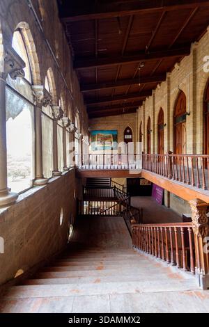 Vista di una scala interna con ringhiere in legno e finestre ad arco, che gettano ombre sulle pareti di pietra, Karachi, Sindh, Pakistan. Foto Stock
