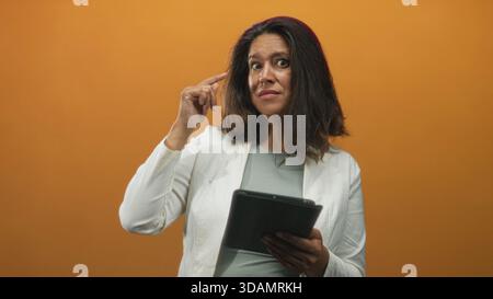 Donna bruna ispanica di mezza età che tiene la tavoletta punta il dito verso il tempio mentre legge e si aggrappa in studio; confusione. Foto Stock