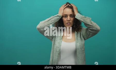 Giovane donna con le mani sulla testa in uno studio di ottanio che indossa canotta bianca e camicia verde chiaro; affaticamento da stress. Foto Stock